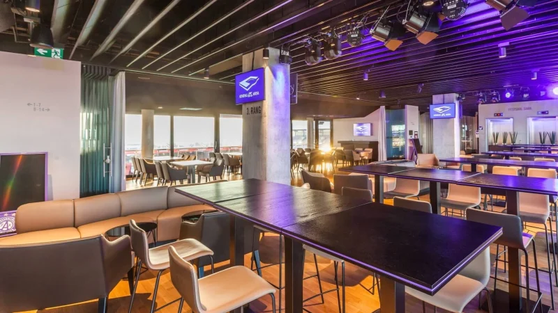 Eine moderne, geräumige Lounge mit hohen Tischen, Barhockern, hellbraunen Ledersesseln, Holzböden und großen Fenstern, die natürliches Licht hereinlassen. Mehrere Bildschirme und Deckenleuchten sorgen für ein modernes Ambiente.