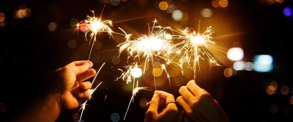 Drei Hände, die Wunderkerzen halten, die in der Nacht hell leuchten, mit einem unscharfen Hintergrund aus Lichtern der Stadt, der den festlichen Geist der Neujahrsvorsätze und der Feierlichkeiten einfängt.