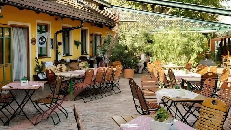Die Terrasse des Restaurants ist mit Holztischen und -stühlen ausgestattet, die teilweise mit karierten Tischtüchern bedeckt sind. Das Gebäude ist gelb mit grünen Verzierungen, und Topfpflanzen schmücken den Bereich und schaffen eine gemütliche und einladende Atmosphäre.