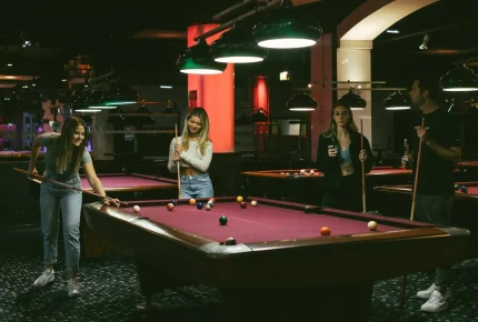 Vier junge Erwachsene spielen in einem schwach beleuchteten Raum um einen lilafarbenen Billardtisch, lächeln und halten Queues. Die Atmosphäre wirkt entspannt und freundlich.