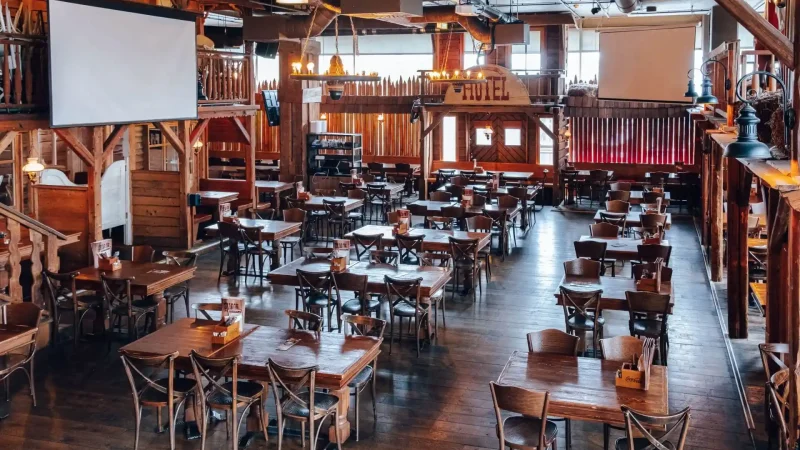 Geräumiges, rustikales Restaurant mit Holztischen und -stühlen, großen Fenstern, Kronleuchtern, zwei Projektionsflächen und einem Balkon. Der Raum ist leer und mit Holzakzenten und Details im westlichen Stil dekoriert.