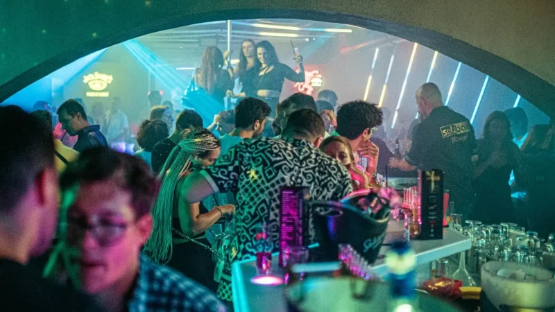 Eine belebte Nachtclubszene mit tanzenden, redenden und trinkenden Menschen unter bunten Neonröhren. Einige Leute stehen an der Bar, während andere auf einer erhöhten Plattform im Hintergrund tanzen und eine lebhafte Partyatmosphäre schaffen.
