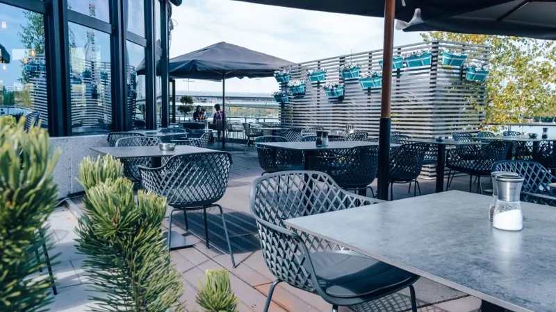 Terrasse eines Außenrestaurants mit modernen Metalltischen und -stühlen, die teilweise von großen Sonnenschirmen beschattet werden. Blaue Blumenkübel hängen an einem Holzlattenzaun. Im Vordergrund stehen Grünpflanzen, und der Bereich scheint menschenleer zu sein.