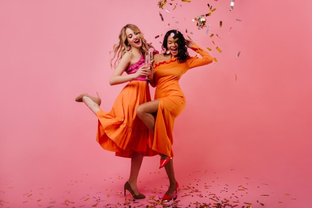 Zwei Frauen in orangefarbenen Kleidern feiern vor einem rosa Hintergrund, lächeln und posieren mit Konfetti in der Luft. Eine hält eine Flasche, beide sehen fröhlich und festlich aus, ganz im Sinne des Partymottos, während um sie herum Konfetti verstreut wird.