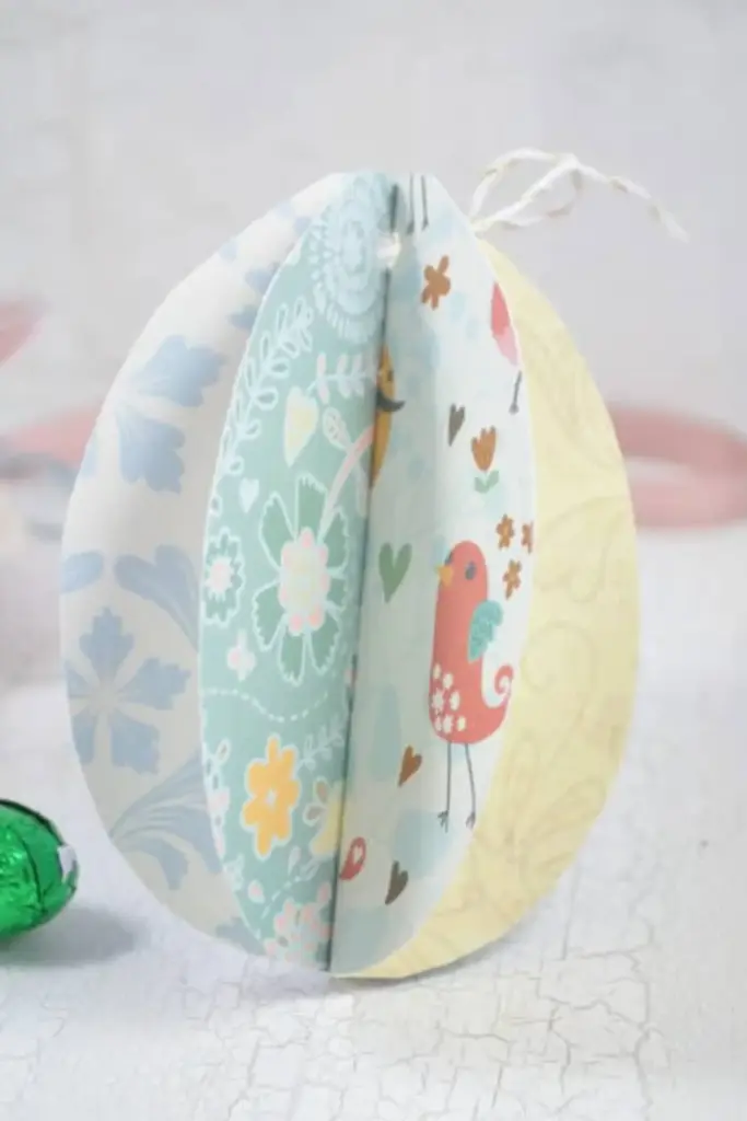 Ein 3D-Papiereiornament aus pastellfarbenem, gemustertem Papier mit Blumen- und Vogelmotiven steht aufrecht mit einer Schnur an der Spitze - perfekt für Osterdeko-Basteln auf einer hellen, strukturierten Oberfläche.