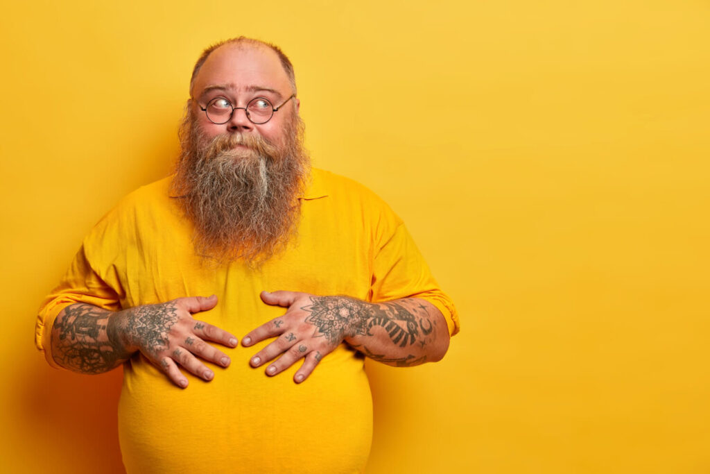 Vor einem gelben Hintergrund steht ein bärtiger Mann mit Brille und Tattoos an den Händen, der ein gelbes Hemd trägt und mit einem verspielten Gesichtsausdruck nach oben schaut - vielleicht träumt er davon, wie er nach den Feiertagen den Weihnachtsspeck loswerden kann.