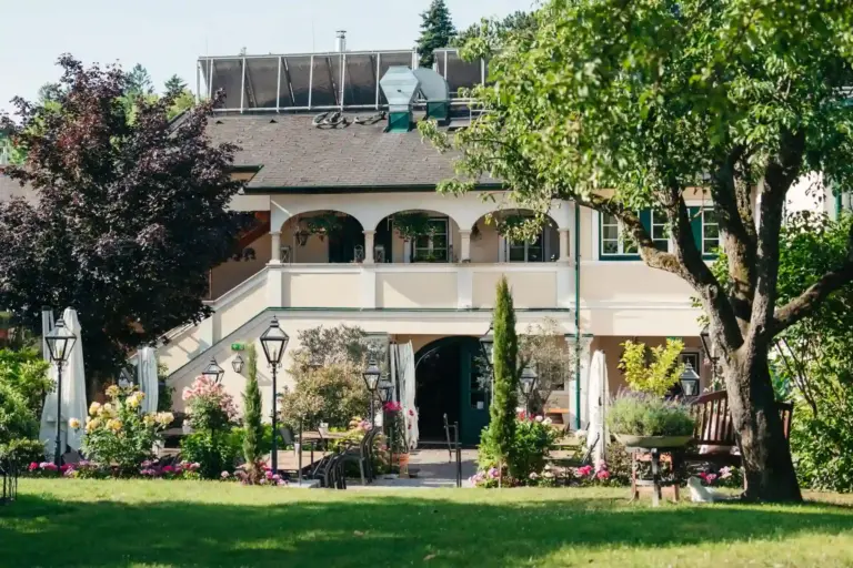 s´Pfiff Restaurant. Ein charmantes, zweistöckiges Gebäude mit gewölbten Balkonen und einem Satteldach, umgeben von üppigem Grün, farbenprächtigen Blumen und Sitzgelegenheiten im Freien auf einem gepflegten Rasen.
