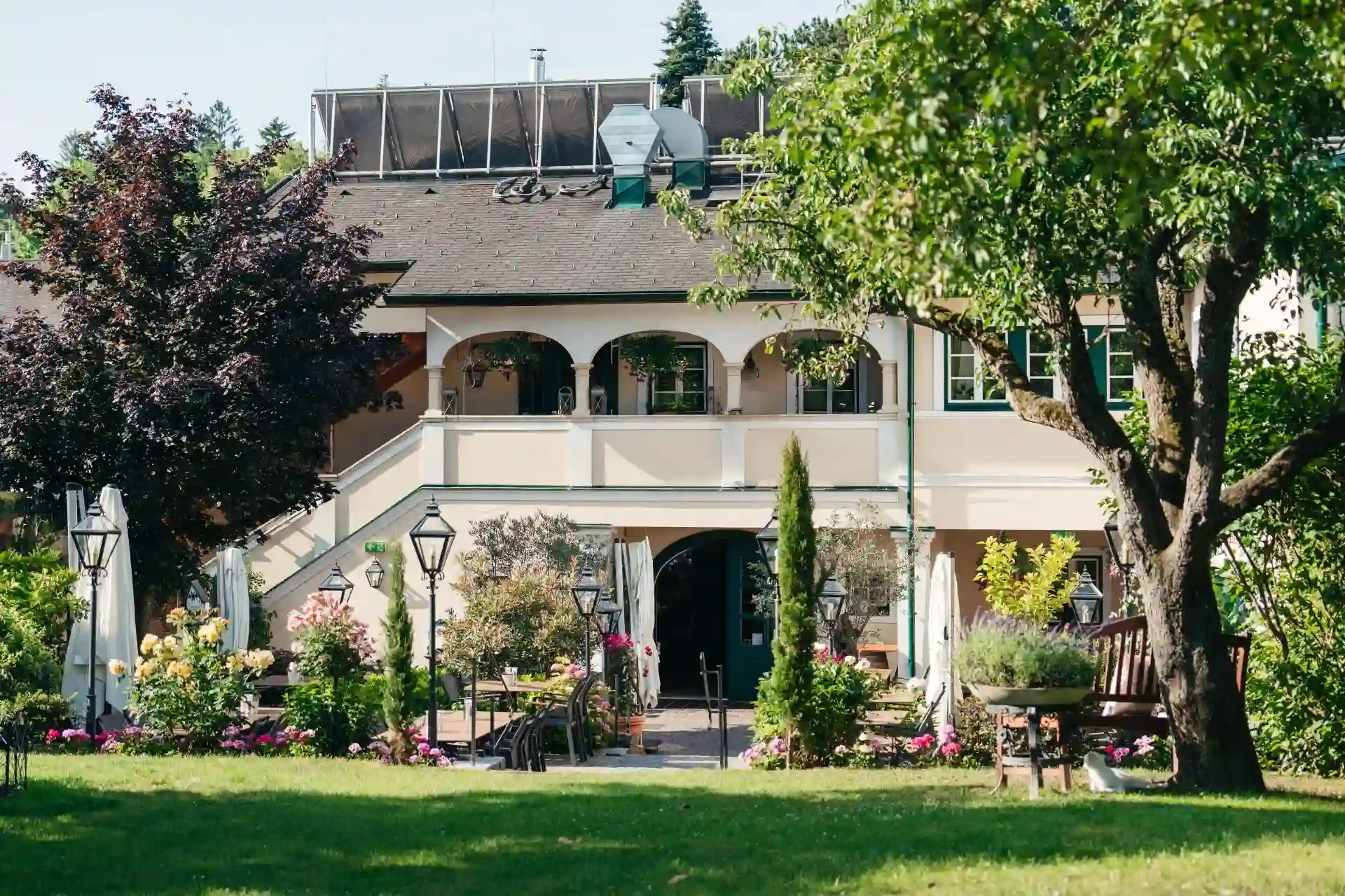 s´Pfiff Restaurant. Ein charmantes, zweistöckiges Gebäude mit gewölbten Balkonen und einem Satteldach, umgeben von üppigem Grün, farbenprächtigen Blumen und Sitzgelegenheiten im Freien auf einem gepflegten Rasen.