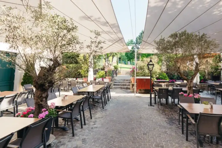 Terrasse eines Restaurants im Freien mit Steinboden, an beiden Seiten aufgestellten Tischen und Stühlen, Sonnensegeln über den Köpfen, Topfpflanzen und Bäumen, die eine gemütliche, einladende Atmosphäre schaffen. Im Hintergrund sind Stufen und Grünflächen zu sehen.