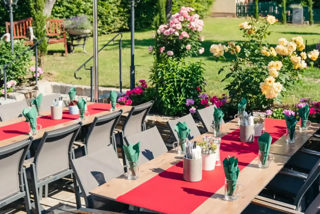 Esstische im Freien mit roten Tischläufern, ordentlich gedeckt mit Gläsern, Besteck und Servietten, umgeben von blühenden Blumen und Grünpflanzen in einer Gartenlandschaft an einem sonnigen Tag.