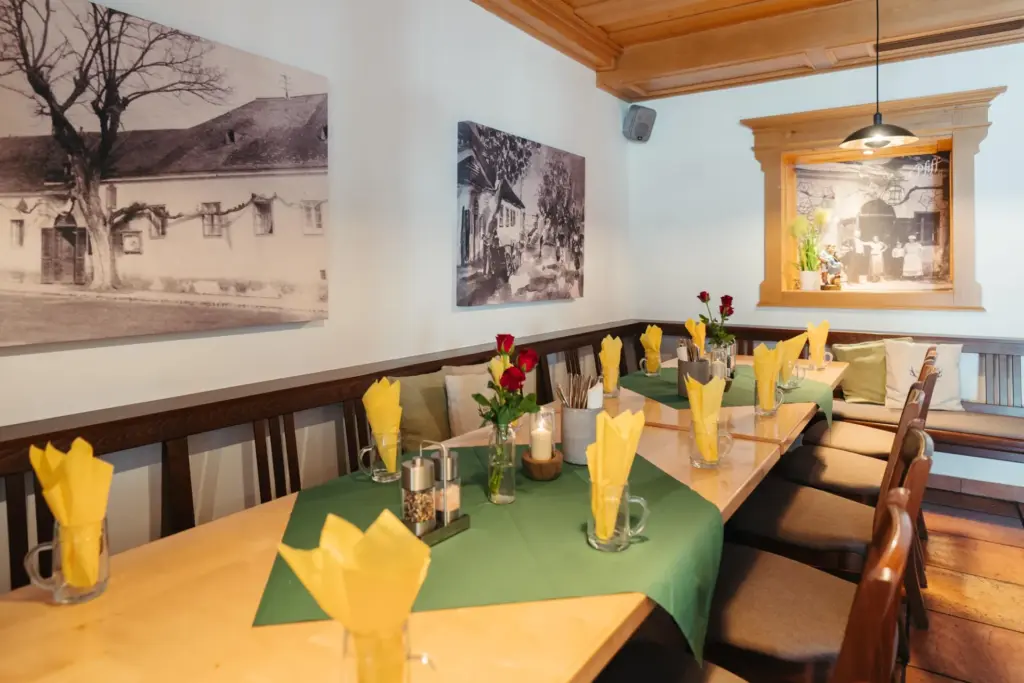 Ein gemütlicher Restaurantbereich mit Holzmöbeln, Tischen, auf denen gelbe Servietten in Glasbechern liegen, grünen Tischläufern und roten Rosen. An den weißen Wänden hängen Schwarz-Weiß-Fotos, und warmes Licht sorgt für eine einladende Atmosphäre.