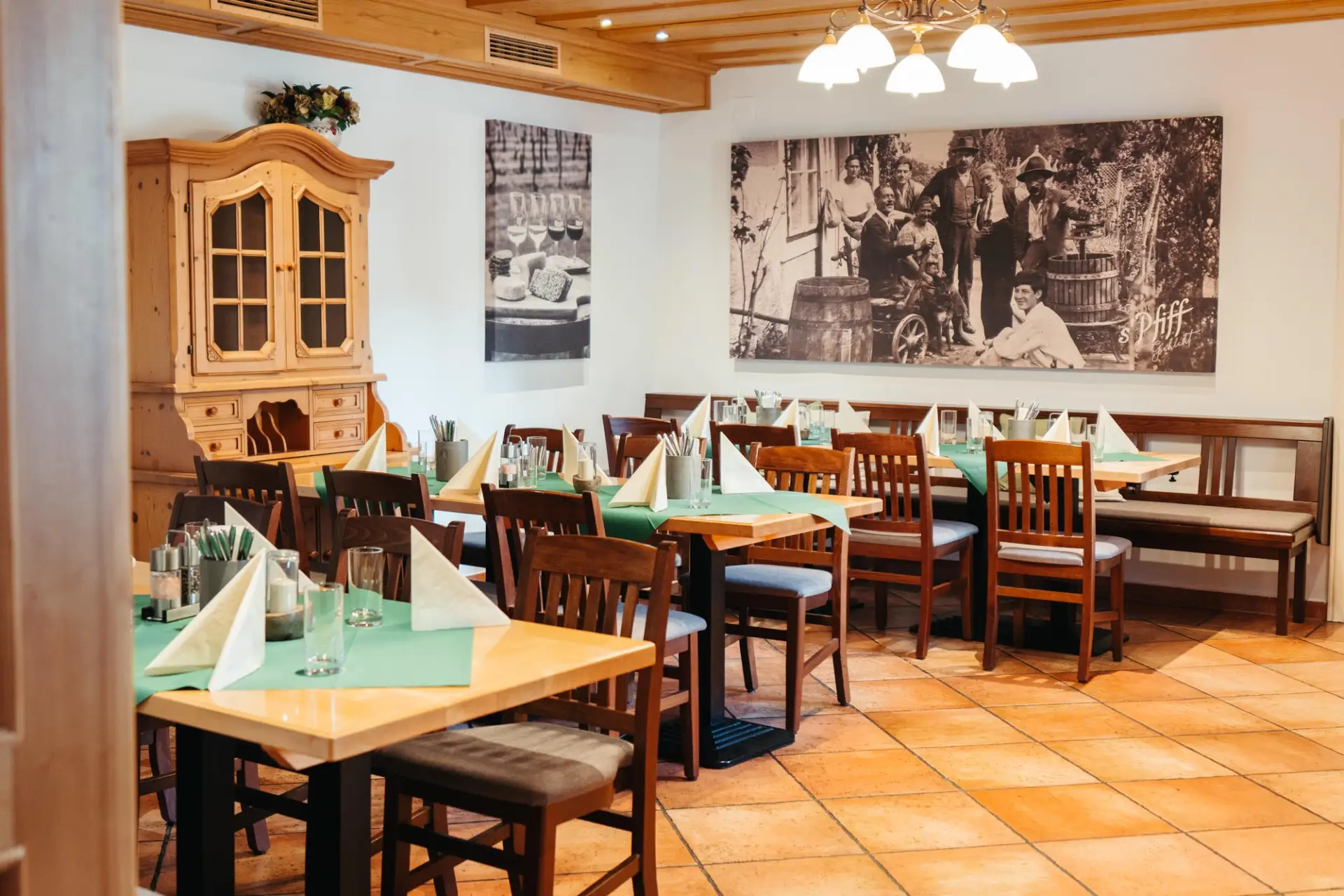 Ein gemütlicher Restaurantbereich mit Holztischen und -stühlen, die für die Gäste gedeckt sind, grünen Tischläufern, Servietten und Gläsern. An den Wänden hängen große Schwarz-Weiß-Fotos und ein Holzschrank. Warme Beleuchtung und ein Fliesenboden sorgen für eine einladende Atmosphäre.