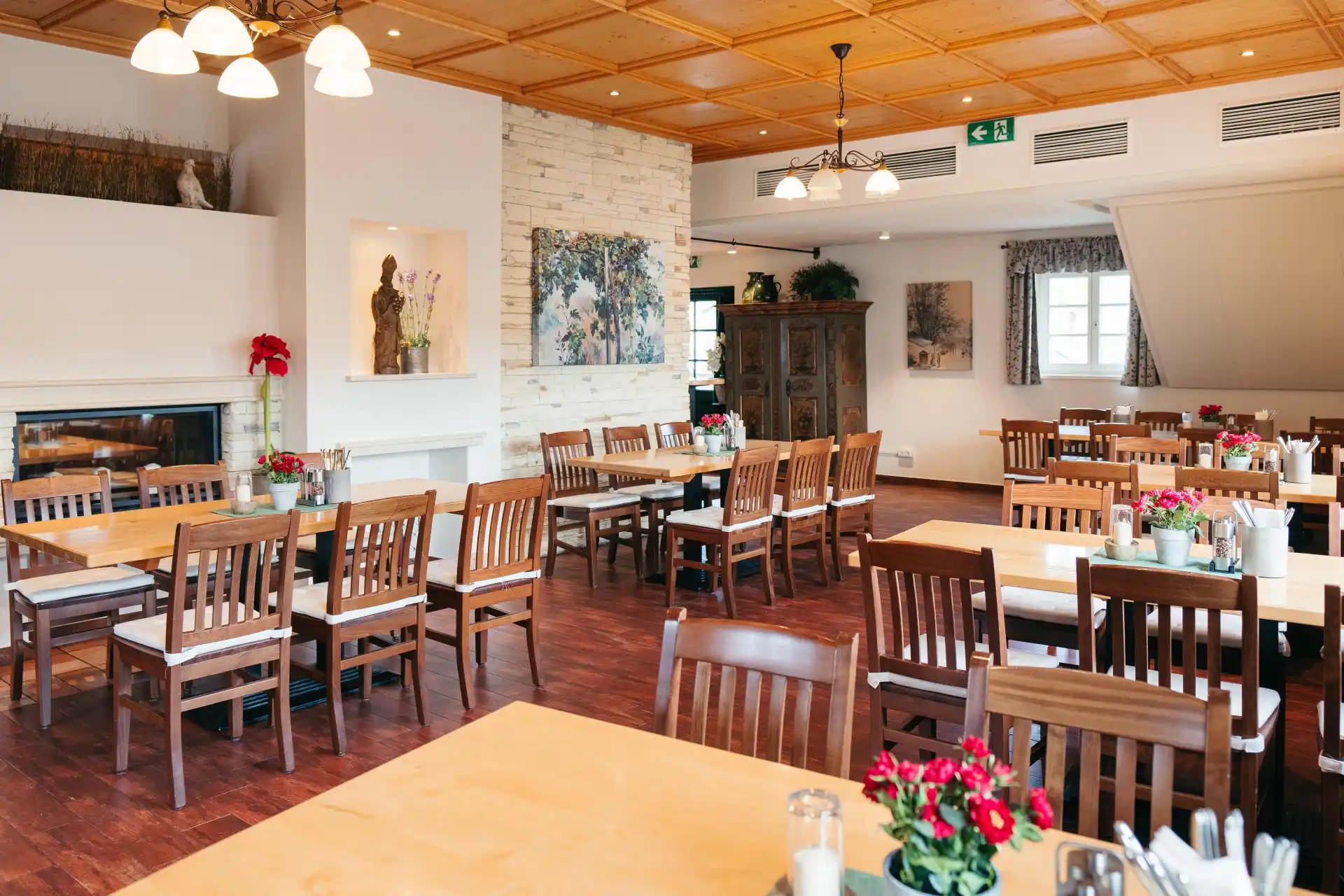 Ein helles, gemütliches Restaurantinterieur mit Holztischen und -stühlen, roten Blumen auf den Tischen, Kunstwerken an den Wänden und warmer Beleuchtung durch Deckenlampen. Der Raum ist menschenleer.