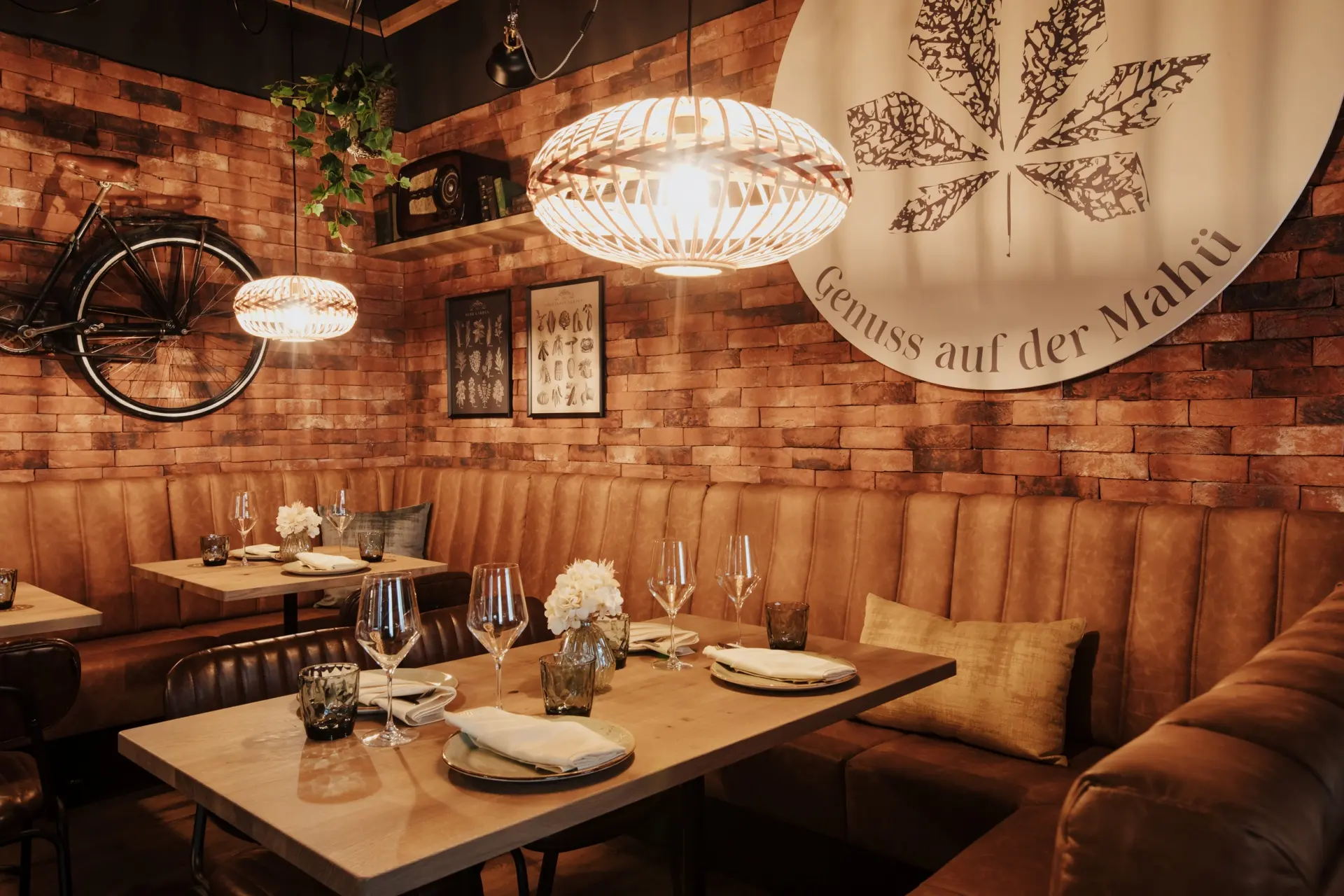 Ein gemütliches Restaurant mit braunen Ledersesseln, Holztischen mit Tellern und Gläsern, Backsteinwänden, warmen Hängelampen, dekorativen Blumen und einem großen Wandbild mit einem Blatt und einem deutschen Text.