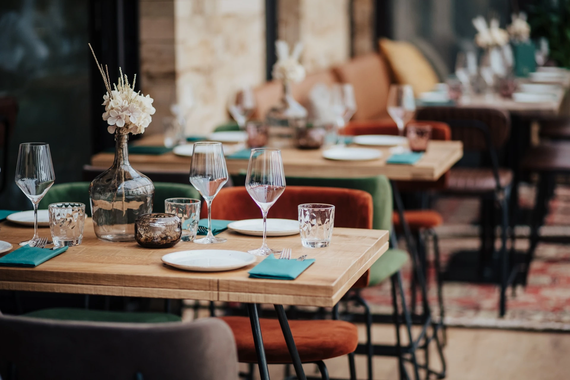 Ein gemütliches Restaurant mit Holztischen, die zum Essen gedeckt sind, mit Tellern, Gläsern, Besteck, blaugrünen Servietten und Blumenvasen. Sanft gepolsterte Stühle umgeben die Tische in einer warm beleuchteten Atmosphäre.