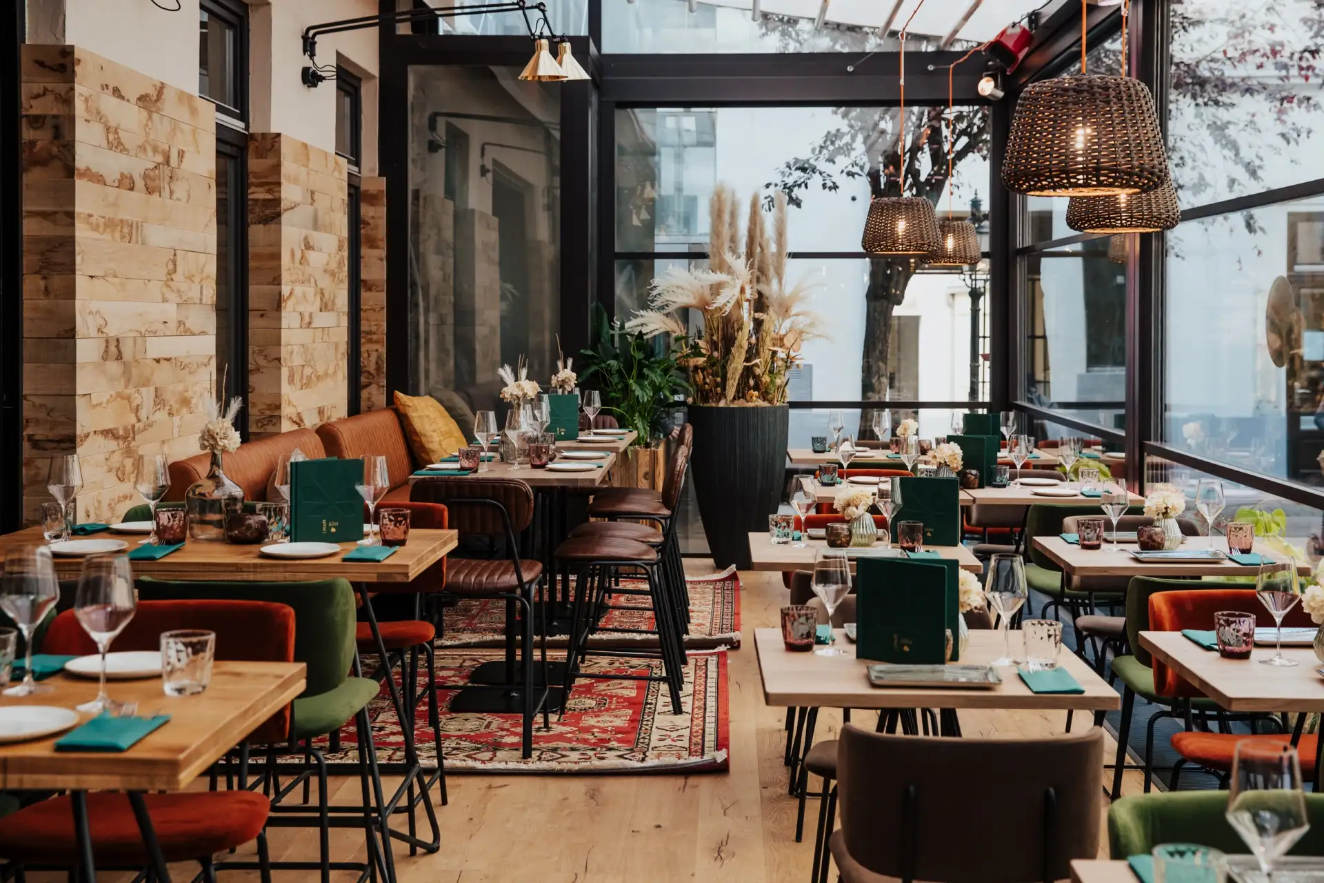Ein modernes Restaurant mit Holztischen, farbenfrohen Stühlen, grün gedeckten Tischen, hängenden Weidenlampen, großen Fenstern und dekorativen Pflanzen, die eine gemütliche und stilvolle Atmosphäre schaffen.