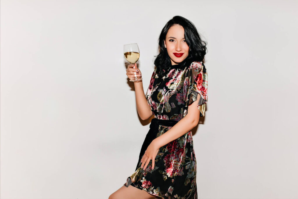 Eine Frau mit gewelltem schwarzem Haar und rotem Lippenstift, die ein geblümtes Kleid trägt, lächelt und hält ein Glas Weißwein in der Hand - was die Atmosphäre einer TOP Location für Weinliebhaber einfängt - vor einem schlichten weißen Hintergrund.