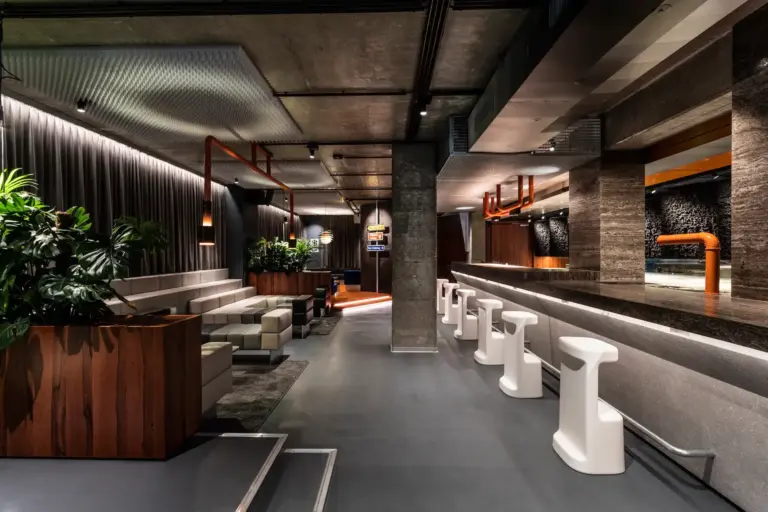 Moderne Lounge-Bar mit glattem grauen Boden, weißen Barhockern, einem langen Bartresen, stilvoller Beleuchtung, Grünpflanzen und modernen Sitzbereichen mit Holz- und Lederakzenten. Die Atmosphäre ist gehoben und einladend.