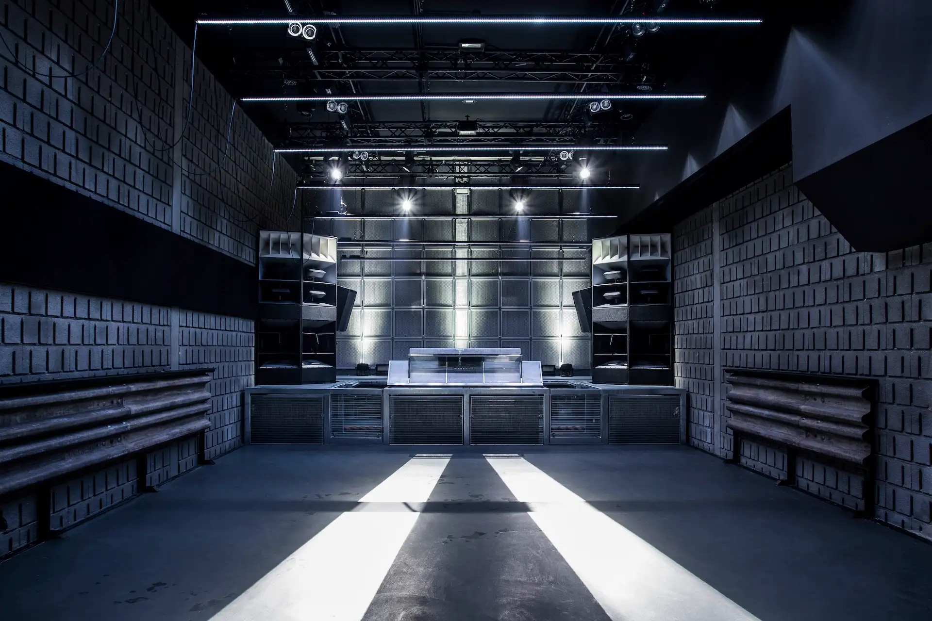 Ein moderner, leerer Nachtclub mit geometrischen, schalldichten Wänden, einem großen DJ-Pult in der Mitte, leistungsstarken Lautsprechern und Lichtstrahlen, die über den Boden auf die minimalistische Bühne scheinen.