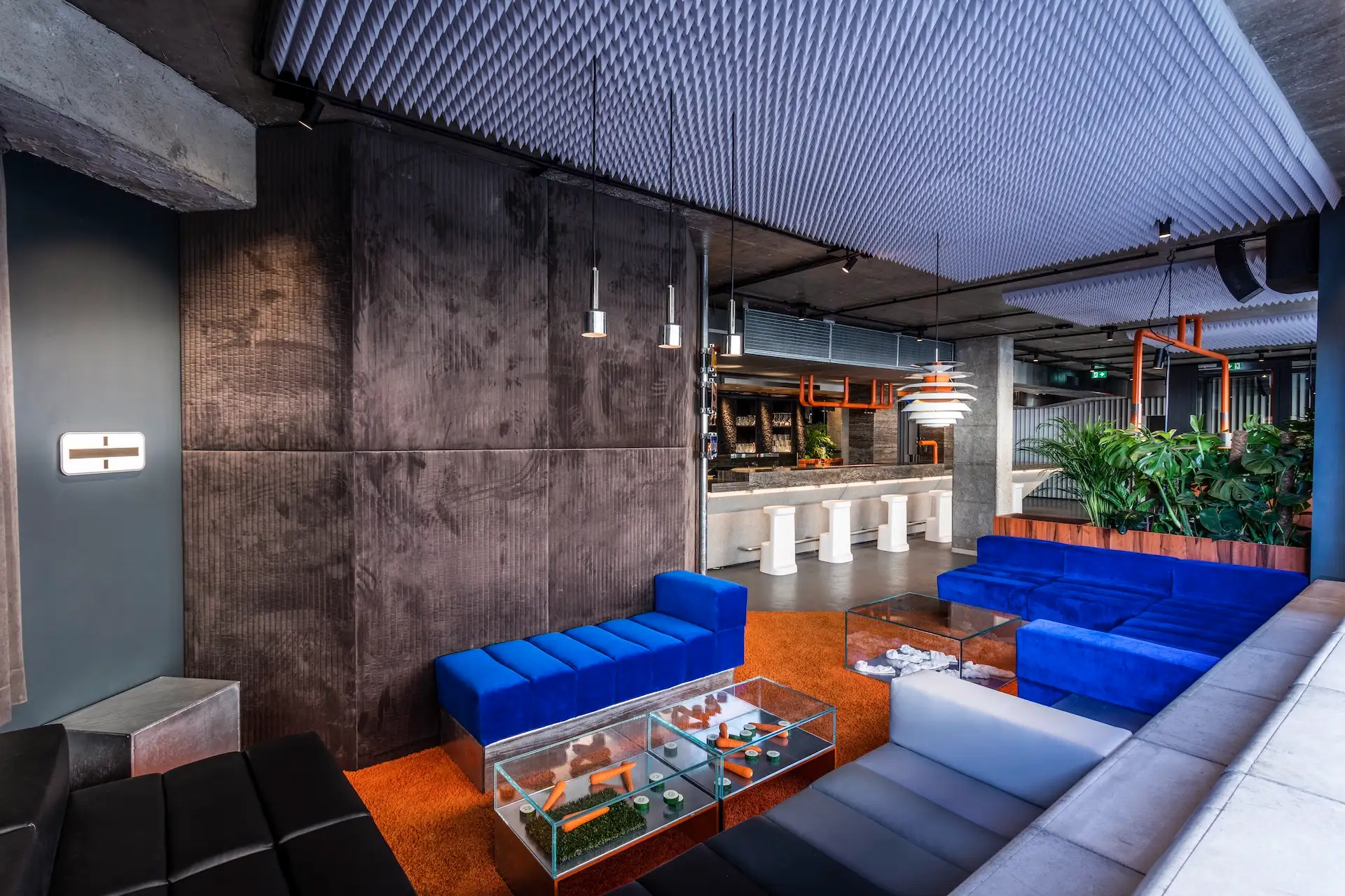 Moderne Lounge mit blauen und weißen Sofas, Couchtischen aus Glas, orangefarbenem Teppich, Hängelampen, Pflanzen und einem Barbereich mit weißen Hockern und einer industriellen Deckengestaltung.