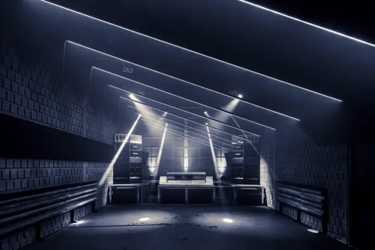 Der moderne, schummrig beleuchtete Nachtclub verfügt über eine zentrale DJ-Kabine, große Lautsprecher und geometrische LED-Lichtstreifen an der Decke, die eine futuristische Atmosphäre schaffen. Die Wände sind mit Akustikplatten verkleidet.